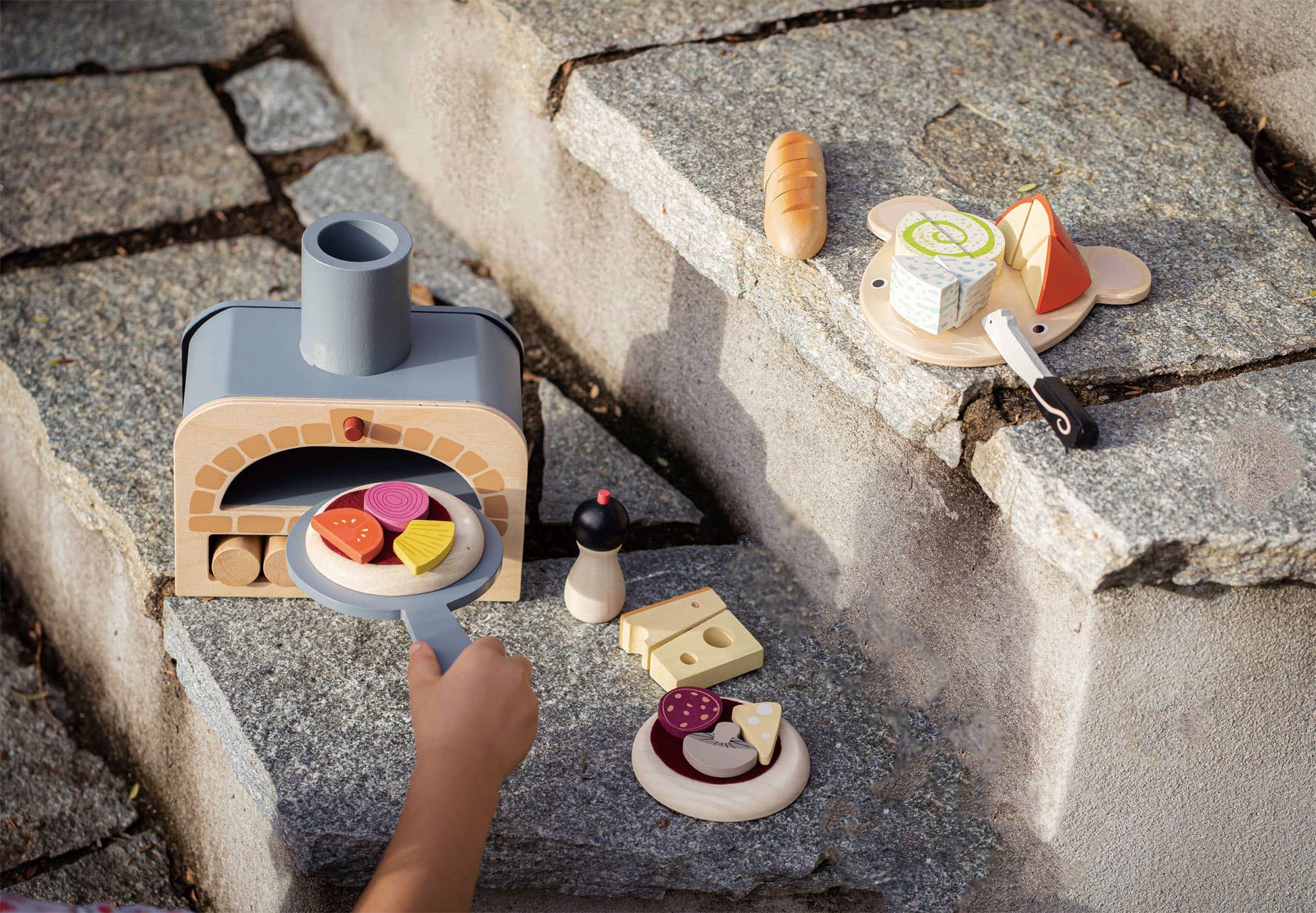 Make Me a Pizza! - Wooden Pizza Oven Toy Play Set in outdoor play scene with additional wooden food items, encouraging creative pretend cooking.