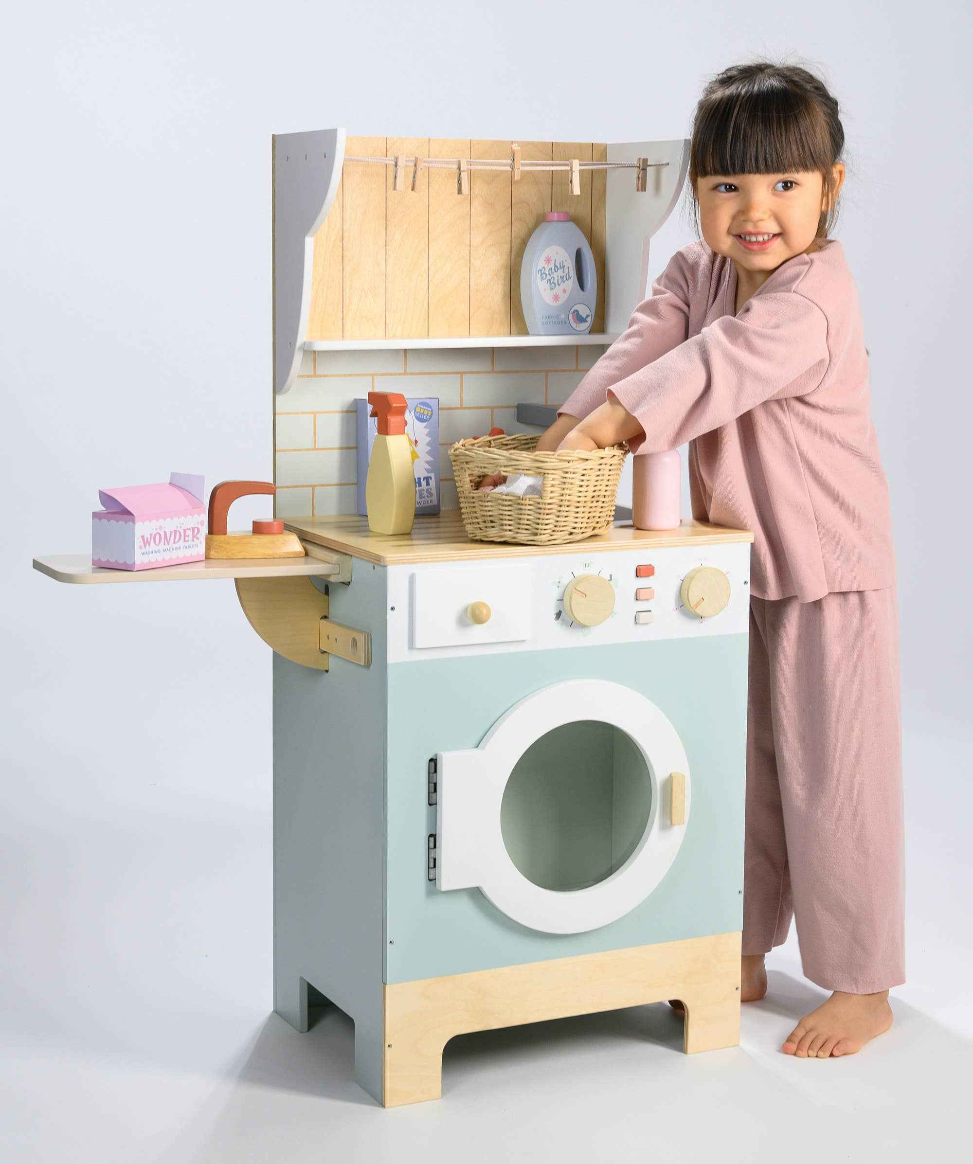 Young girl using Tender Leaf Wooden Laundrette, placing items into a basket on the toy laundry station for role play fun.