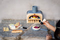 Child using Make Me a Pizza! - Wooden Pizza Oven Toy Play Set outdoors, adding toppings and engaging in imaginative cooking role play.